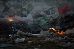 A pile of garbage is burning, with flames visible among the trash which includes plastic bags and various debris. Smoke rises, creating a hazy atmosphere.