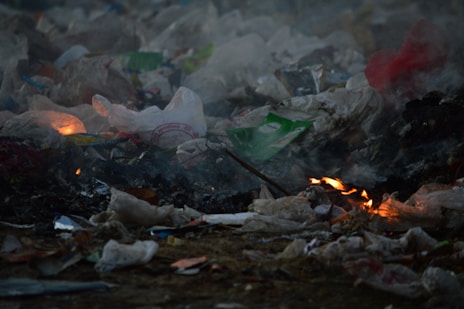 A pile of garbage is burning, with flames visible among the trash which includes plastic bags and various debris. Smoke rises, creating a hazy atmosphere.