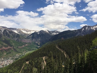 A scenic mountain landscape with snow-capped peaks under a partially cloudy blue sky. Dense green forests cover the slopes and valleys, with a small town nestled among the greenery. Dirt paths wind through the forest, and ski lift chairs are visible.