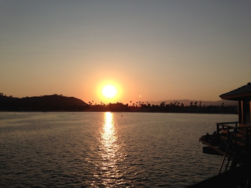 A serene sunset over a large body of water with distant palm trees and hills silhouetted against the golden sky. A structure with a wooden deck is visible on the right side, suggesting a peaceful waterfront setting.