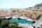 A coastal cityscape featuring numerous modern buildings along the waterfront. Luxurious yachts are lined up in a marina, with a backdrop of hills and the ocean. The architecture displays varied colors and styles, reflecting a vibrant, upscale area.