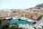 A coastal cityscape featuring numerous modern buildings along the waterfront. Luxurious yachts are lined up in a marina, with a backdrop of hills and the ocean. The architecture displays varied colors and styles, reflecting a vibrant, upscale area.