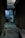 A dimly lit alleyway lined with closed shutters leads to a shop. A signboard reads 'Raja Tailors Only for Ladies Tailors'. Inside, a person is visible, working on something at a table surrounded by various objects.