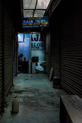 A dimly lit alleyway lined with closed shutters leads to a shop. A signboard reads 'Raja Tailors Only for Ladies Tailors'. Inside, a person is visible, working on something at a table surrounded by various objects.