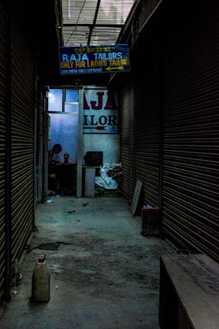A dimly lit alleyway lined with closed shutters leads to a shop. A signboard reads 'Raja Tailors Only for Ladies Tailors'. Inside, a person is visible, working on something at a table surrounded by various objects.