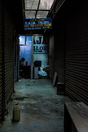 A dimly lit alleyway lined with closed shutters leads to a shop. A signboard reads 'Raja Tailors Only for Ladies Tailors'. Inside, a person is visible, working on something at a table surrounded by various objects.