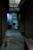 A dimly lit alleyway lined with closed shutters leads to a shop. A signboard reads 'Raja Tailors Only for Ladies Tailors'. Inside, a person is visible, working on something at a table surrounded by various objects.