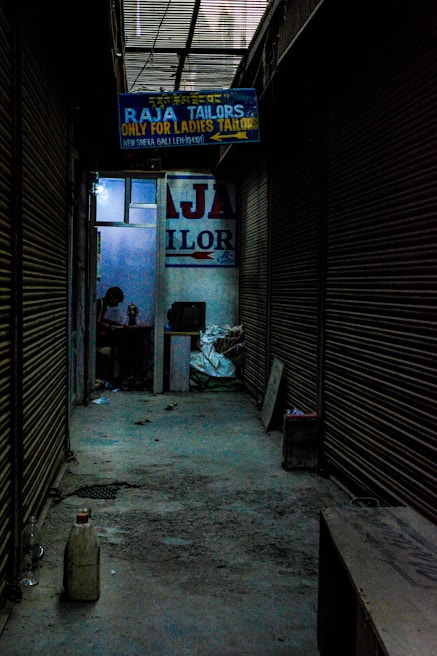 A dimly lit alleyway lined with closed shutters leads to a shop. A signboard reads 'Raja Tailors Only for Ladies Tailors'. Inside, a person is visible, working on something at a table surrounded by various objects.