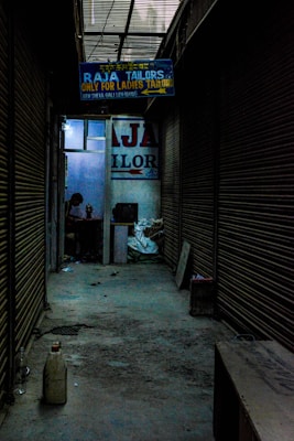 A dimly lit alleyway lined with closed shutters leads to a shop. A signboard reads 'Raja Tailors Only for Ladies Tailors'. Inside, a person is visible, working on something at a table surrounded by various objects.