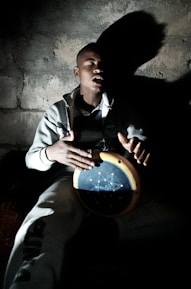 A man with closed eyes is playing a handheld drum in a dimly lit environment, casting a prominent shadow against a textured wall. He wears a casual grey tracksuit and appears to be deeply absorbed in his music.