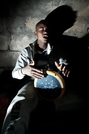 A man with closed eyes is playing a handheld drum in a dimly lit environment, casting a prominent shadow against a textured wall. He wears a casual grey tracksuit and appears to be deeply absorbed in his music.