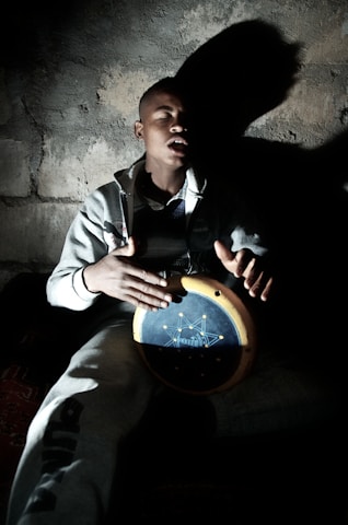 A man with closed eyes is playing a handheld drum in a dimly lit environment, casting a prominent shadow against a textured wall. He wears a casual grey tracksuit and appears to be deeply absorbed in his music.