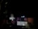 A dimly lit area with a dark background is visible. There is a central object resembling a security camera or electronic device. Below it, there is a rectangular white box with some graphics or text on its side. Cables or wires appear to be hanging or connected among the items.