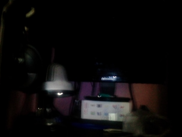 A dimly lit area with a dark background is visible. There is a central object resembling a security camera or electronic device. Below it, there is a rectangular white box with some graphics or text on its side. Cables or wires appear to be hanging or connected among the items.