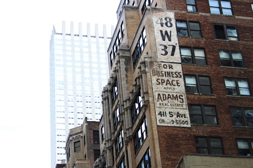 A juxtaposition of an older, brick office building with a large advertisement for business space, and a modern, sleek glass skyscraper in the background. The older building features multiple windows and a vintage sign promoting Adams & Co. Real Estate at 411 5th Ave.