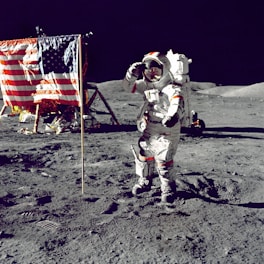 An astronaut in a white spacesuit stands on a barren lunar surface near a U.S. flag, saluting. The background includes a lunar lander and grey rocky terrain under a black sky.