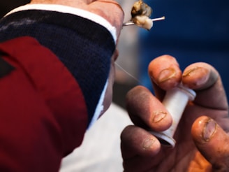 A close-up view of a person's hands engaged in threading. One hand holds a needle with a piece of thread, while the other grips a spool of thread. The hands appear rough and slightly dirty, suggesting they belong to someone who works with their hands regularly. The background is blurred, emphasizing the focus on the threading activity.