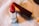 A red lipstick with a shiny silver casing is positioned on a soft, blurred background. The lipstick is partially extended, showing its vibrant color with a nearby closed cap.