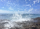 A powerful ocean wave crashes against rocky shorelines, sending an explosion of white foam and water droplets into the air. Clear blue skies stretch above, creating a vivid contrast with the deep blue of the sea. The energy of the water is captured in the mid-splash, with large and small droplets scattered across the scene.