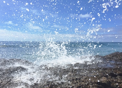 A powerful ocean wave crashes against rocky shorelines, sending an explosion of white foam and water droplets into the air. Clear blue skies stretch above, creating a vivid contrast with the deep blue of the sea. The energy of the water is captured in the mid-splash, with large and small droplets scattered across the scene.