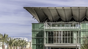 A modern building with a large glass facade and distinctive overhanging roof panels. The text 'Dr. Phillips Center' is visible in white letters across the front. Palm trees are in the foreground, suggesting a warm climate. The sky is clear with some wispy clouds.