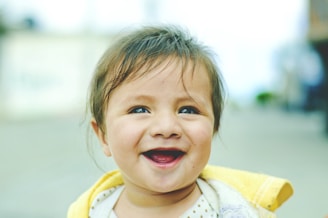 A young child with light brown hair is smiling joyfully outdoors. The child wears a yellow and white outfit, and their expression is one of happiness and contentment against a blurred background.