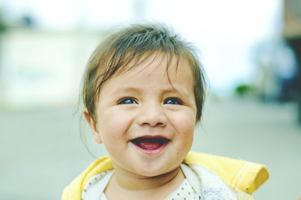 A young child with light brown hair is smiling joyfully outdoors. The child wears a yellow and white outfit, and their expression is one of happiness and contentment against a blurred background.