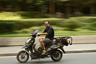 A man riding a motor scooter on a road, with a motion blur effect capturing the sense of speed. He is wearing a black shirt and gray shorts. The scooter carries a metal cylinder and appears to be adapted for carrying cargo. The background is a green, natural setting.