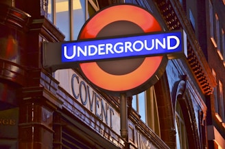 A red and blue sign with the word 'UNDERGROUND' is prominently displayed, attached to a brick wall that has reflective surfaces. The architecture is classic, and there are illuminated windows in the background.