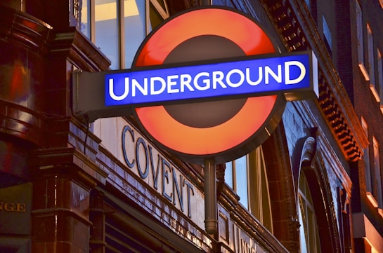 A red and blue sign with the word 'UNDERGROUND' is prominently displayed, attached to a brick wall that has reflective surfaces. The architecture is classic, and there are illuminated windows in the background.