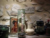 A rustic kitchen setting with a rough stone wall backdrop. Two glass jars with vintage labels are filled with grains and flours, standing next to a white ceramic teapot with floral patterns. Several ceramic containers, including a dark green jar and a cream-colored pot, are arranged alongside a small metal scoop.