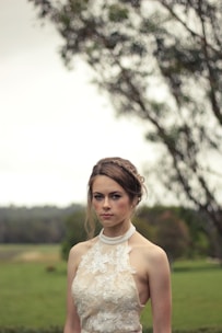 A woman with braided hair is wearing a sleeveless lace dress and standing outdoors. The background features blurred greenery and large trees, giving a serene and natural setting.