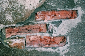 An exposed portion of a weathered brick wall partially covered by concrete and sand. The bricks have a reddish-brown coloration with rough textures, surrounded by grayish cement and patches of dirt.
