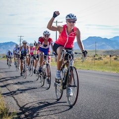 A group of cyclists is riding on a rural road with mountains in the background. The leading cyclist, wearing a pink jersey and sunglasses, raises an arm in triumph or excitement. Other cyclists are following closely, some with smiles and positive gestures. The scene is sunlit with a clear sky and open landscape.
