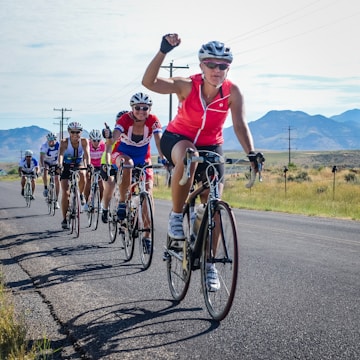 A group of cyclists is riding on a rural road with mountains in the background. The leading cyclist, wearing a pink jersey and sunglasses, raises an arm in triumph or excitement. Other cyclists are following closely, some with smiles and positive gestures. The scene is sunlit with a clear sky and open landscape.