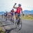 A group of cyclists is riding on a rural road with mountains in the background. The leading cyclist, wearing a pink jersey and sunglasses, raises an arm in triumph or excitement. Other cyclists are following closely, some with smiles and positive gestures. The scene is sunlit with a clear sky and open landscape.