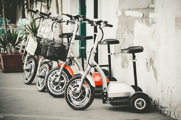A lineup of four scooters parked against a textured wall. The scooters feature large wheels and handlebars, some with baskets. The setting appears urban, with plants in pots and worn concrete surfaces around.