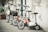 A lineup of four scooters parked against a textured wall. The scooters feature large wheels and handlebars, some with baskets. The setting appears urban, with plants in pots and worn concrete surfaces around.