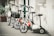 A lineup of four scooters parked against a textured wall. The scooters feature large wheels and handlebars, some with baskets. The setting appears urban, with plants in pots and worn concrete surfaces around.