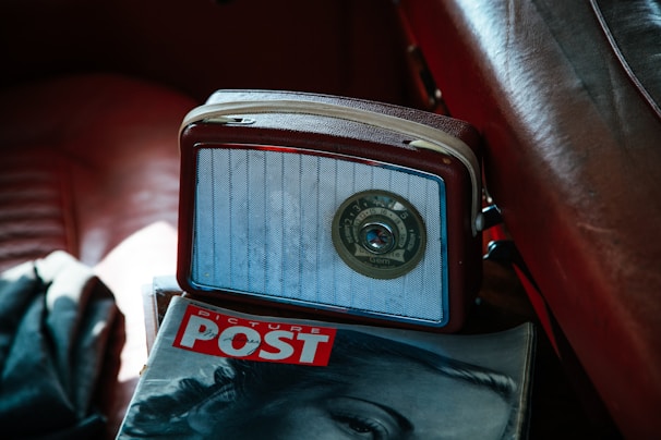 A vintage radio with a central tuning dial rests on a leather seat. Next to it lies a magazine with a partial cover image of a woman's face and the title 'PICTURE POST' in bold red letters, creating a nostalgic ambiance.