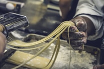 A person is using a pasta maker to cut freshly rolled dough into strips of pasta. The hands are dusted with flour, and the pasta sheets are being guided through the machine. The setting appears to be a kitchen, with a focus on the process of making homemade pasta.