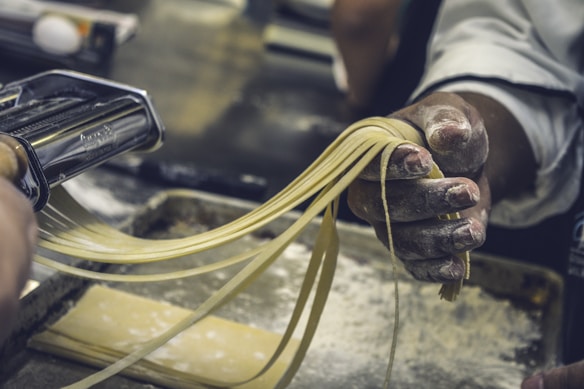 A person is using a pasta maker to cut freshly rolled dough into strips of pasta. The hands are dusted with flour, and the pasta sheets are being guided through the machine. The setting appears to be a kitchen, with a focus on the process of making homemade pasta.