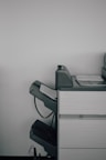 A large office printer or copier is positioned against a plain white wall. The machine is gray with a few sheets of paper visible in the output tray, and there are multiple compartments and trays, suggesting functionality for various paper sizes and types.