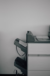 A large office printer or copier is positioned against a plain white wall. The machine is gray with a few sheets of paper visible in the output tray, and there are multiple compartments and trays, suggesting functionality for various paper sizes and types.