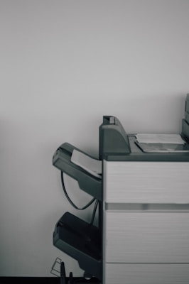 A large office printer or copier is positioned against a plain white wall. The machine is gray with a few sheets of paper visible in the output tray, and there are multiple compartments and trays, suggesting functionality for various paper sizes and types.