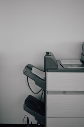 A large office printer or copier is positioned against a plain white wall. The machine is gray with a few sheets of paper visible in the output tray, and there are multiple compartments and trays, suggesting functionality for various paper sizes and types.