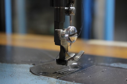 A close-up of a sewing machine needle and presser foot, capturing the metallic components and intricate details. The background is slightly blurred, emphasizing the focus on the sewing mechanism.