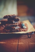 A stack of chocolate brownies is arranged on a piece of parchment paper resting on a wire rack. The brownies have a rich and fudgy texture with a slightly cracked top, suggesting they're freshly baked. The background is softly blurred, drawing focus to the dessert.