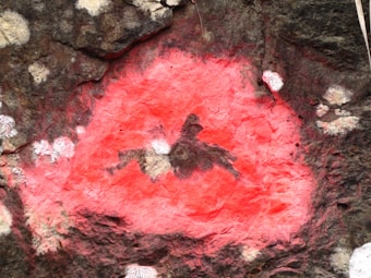 A rock surface featuring a striking red pigment, with abstract shapes and patterns creating a central motif. The surrounding rock has a rough, textured appearance with natural beige and dark brown tones.