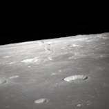 A barren, grey lunar landscape with multiple craters of varying sizes scattered across the surface. A distinct meandering line runs toward the horizon, possibly indicating a ridge or fault line. The dark sky above provides stark contrast to the illuminated surface.